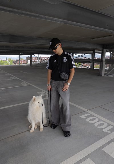 Una persona con una camiseta de polo negra, pantalones de pierna ancha grises y zapatos negros está de pie junto a un perro Samoyedo blanco en una estructura de estacionamiento de concreto.