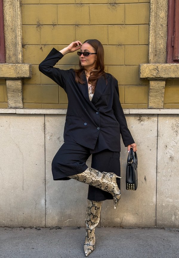 Mujer con blazer negro oversize y pantalones, botas beige con estampado de serpiente, gafas de sol, sosteniendo un bolso negro, posando frente a una pared amarilla y beige.