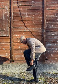 Un homme portant une casquette et un blazer se penche pour ajuster une botte en cuir noir devant un mur en bois usé en plein air.