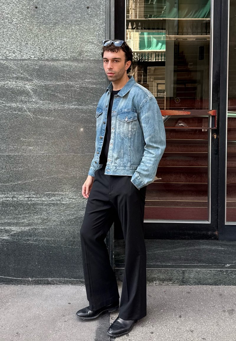 Uomo che indossa una giacca di jeans azzurro chiaro, una camicia nera, pantaloni larghi neri e scarpe nere, si trova con una mano in tasca accanto a una porta di vetro.