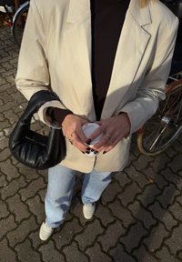 Roomkleurig colbert, zwarte geribbelde coltrui, lichtblauwe spijkerbroek en witte sneakers. Houdt een koffiebeker vast, met een zwarte hobo-tas en zilveren ringen.
