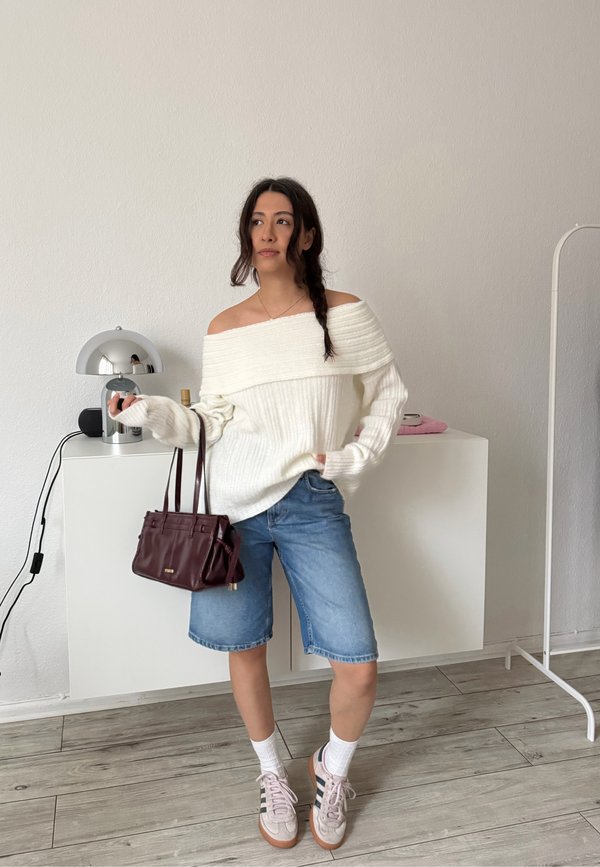 Ženska v belem puloverju brez ramen, jeansa kratkih hlačah, belih nogavicah in supergah, ki drži temno rjavo torbico, stoji v minimalistični sobi.