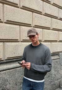 Ung man i grå tröja och mössa som använder smartphone, står mot en strukturerad stenvägg utomhus.