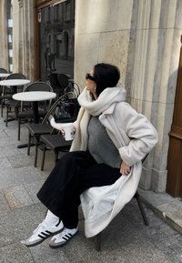 Biely plyšový kabát cez sivý sveter, čierne široké nohavice a šedobiele športové topánky s čiernymi pruhmi. Čierna kabelka, ktorá sedí na stoličke.