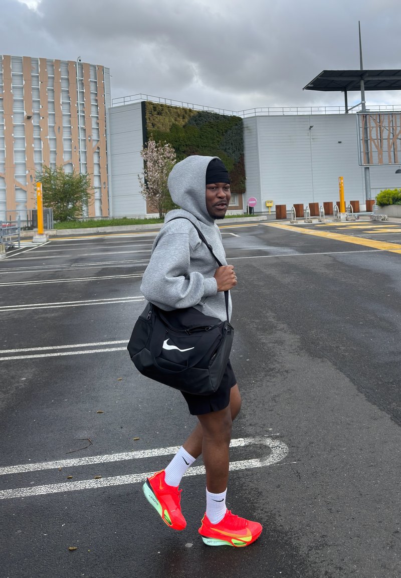 Άτομο με γκρι φούτερ με κουκούλα, μαύρες σορτς και φωτεινά κόκκινα αθλητικά παπούτσια περπατά σε άδειο χώρο στάθμευσης κρατώντας μια μαύρη τσάντα Nike στον έναν ώμο.
