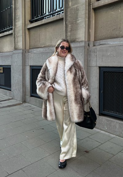 Mujer con abrigo de piel, suéter blanco de punto grueso, pantalones de satén crema, zapatos negros, llevando un bolso negro, caminando por la acera junto a un edificio de piedra.
