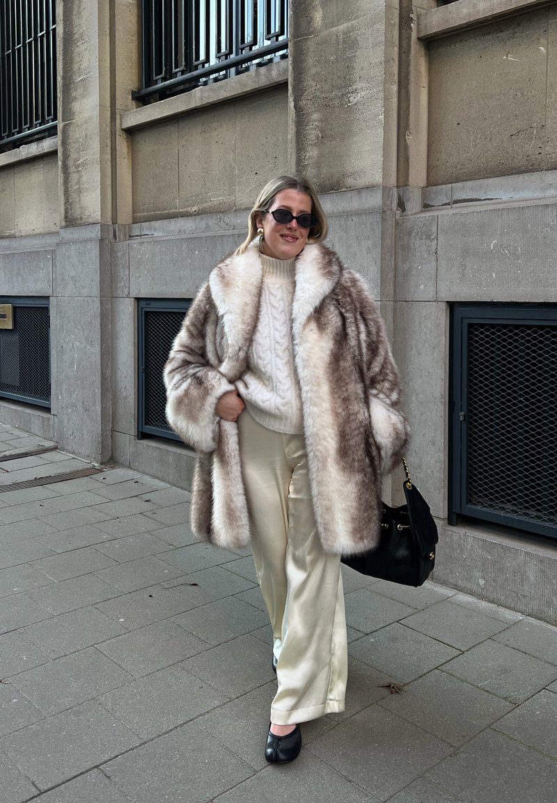 Femme portant un manteau en fourrure, un pull en maille torsadée blanc, un pantalon en satin crème, des chaussures noires, tenant un sac à main noir, marchant sur le trottoir à côté d'un bâtiment en pierre.