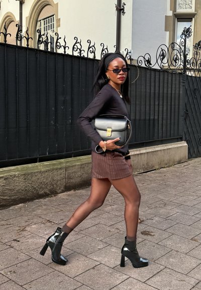 Top negro de manga larga, falda mini con rayas marrones y botas de tacón alto negras y brillantes. Bolso de hombro con detalles dorados y gafas de sol.