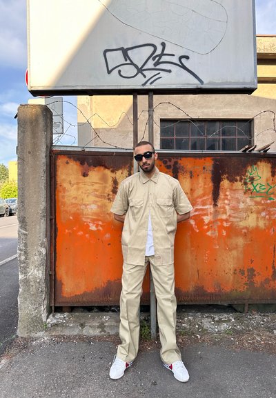 Camisa beige de manga corta y pantalones a juego con dos bolsillos delanteros, combinados con zapatillas blancas. El fondo presenta una superficie de óxido naranja.