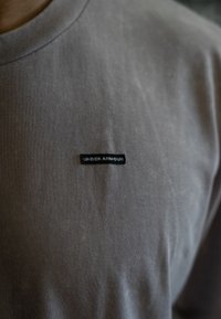 T-shirt cinza de algodão com gola redonda e pequena etiqueta preta "UNDER ARMOUR" no lado esquerdo do peito. Textura suave.
