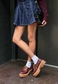 Persoană purtând o fustă albastră, pulover marsala, șosete albe și pantofi platformă maro groși, stând cu picioarele încrucișate lângă o ușă neagră.