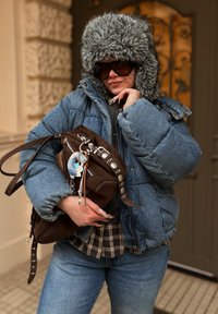 Džínová péřová bunda, šedá kapuce lemovan�á umělou kožešinou, černý svetr, kostkovaná košile, hnědá kabelka se stříbrnými kovovými doplňky a modrý přívěsek jako ozdoba.