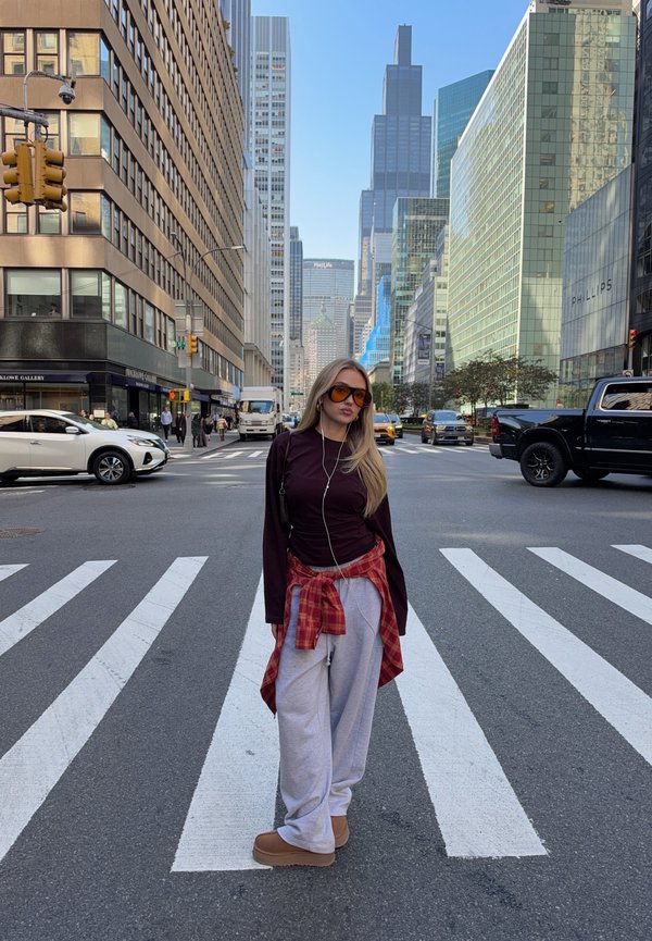Une femme portant des lunettes de soleil pose sur un passage piéton en ville, au milieu de la circulation et de hauts immeubles de bureaux.