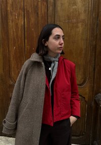 Jeune femme aux cheveux courts et foncés portant une veste rouge sur un haut noir et une écharpe grise, debout devant une porte en bois.