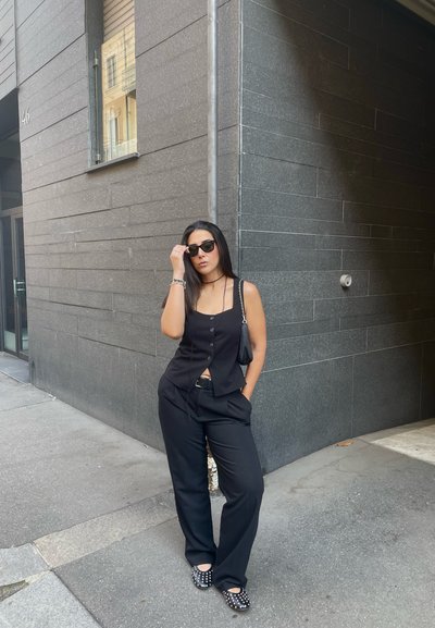 Camiseta tipo chaleco negra de botones, combinada con pantalones negros holgados. Detalles destacables incluyen zapatos negros planos con tachuelas plateadas y un pequeño bolso negro.