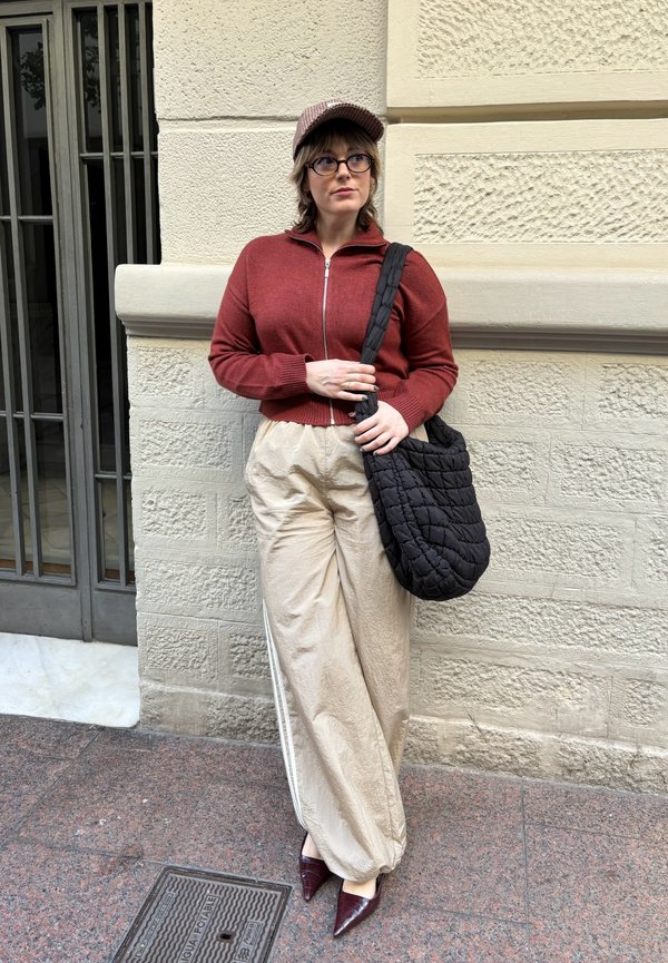 Rød lynlås-sweater, beige bredbenede bukser, sort quilted taske og burgunder spidse hæle. En brun mønstret kasket og briller fuldender looket.