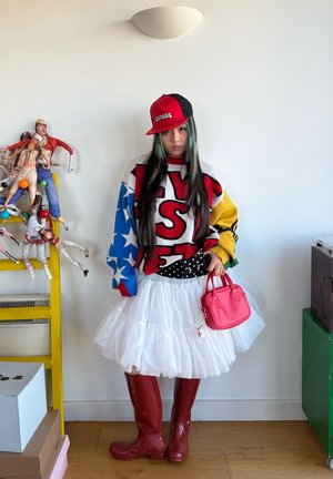 Persona joven vistiendo un suéter colorido y holgado, falda blanca con volantes, botas rojas, gorra roja y negra, sosteniendo un bolso pequeño rosa en el interior.