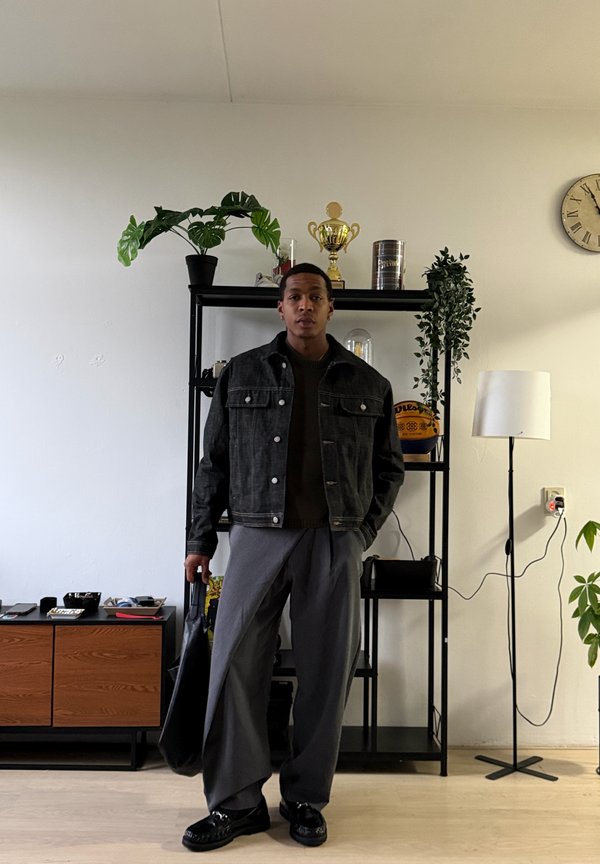 Man draagt een donkere jas en grijze broek, staat binnen en houdt een zwarte tas vast, met een plank, planten, een trofee, een basketbal en een vloerlamp achter hem.