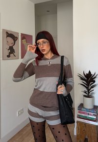 Kvinde med rødt hår justerer briller, iført stribede shorts og sweater, sorte polka-dot strømpebukser, holder en sort taske i et rum med kunst og en plante.