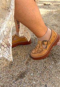 Pantofi din piele maro cu cataramă, design împletit și tălpi groase din cauciuc maro. Purtați cu șosete scurte bej deschis pe un fundal nisipos.