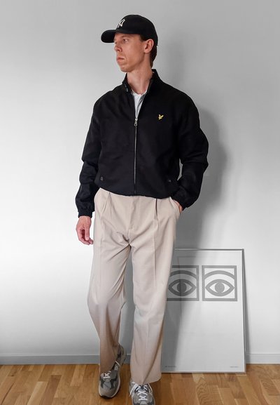 Chaqueta negra con cremallera y puños elásticos, pantalones beiges de pliegues y zapatillas grises. Expuestos contra un fondo minimalista con obras de arte.