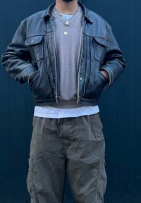 Fekete bőrdzseki két elülső zsebbel, szürke kötött pulóverral alatta, fehér inggel rétegezve, és olívazöld katonai nadrággal.