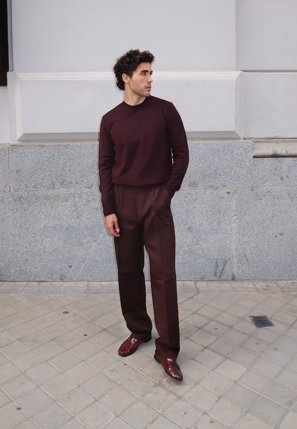 Burgundowy sweter i pasujące spodnie, gładka faktura. Model stoi przy szarej kamiennej ścianie, nosząc ciemne buty ze spiczastym noskiem.