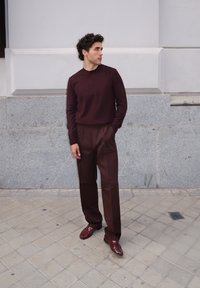 Burgundowy sweter i pasujące spodnie, gładka faktura. Model stoi przy szarej kamiennej ścianie, nosząc ciemne buty ze spiczastym noskiem.