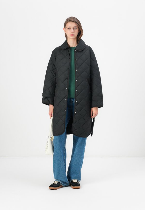 Schwarzer steppgeführter langer Mantel mit Kragen, Druckknöpfen und Seitentaschen. Gestylt über einem grünen Pullover und kombiniert mit weitgeschnittenen blauen Jeans und Sneakers.