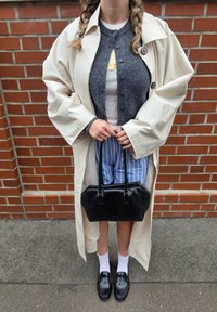 Beige trench coat, grå cardigan, hvit t-skjorte, blåstripete shorts, svart håndveske, hvite sokker og svarte sko. Mursteinsbakgrunn.