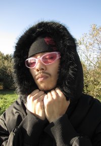 Casaco preto com capuz forrado a pêlo, gorro preto e óculos de sol cor-de-rosa. O tecido é liso; o pêlo adiciona textura. Mãos entrelaçadas sob o queixo.