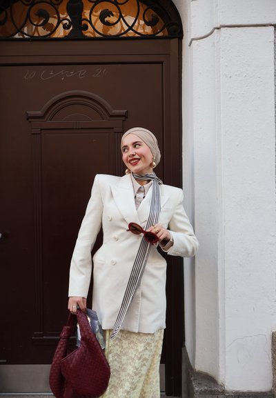 Blazer blanco de doble botonadura, bufanda a rayas, hiyab beige. Sosteniendo gafas de sol rojas y un bolso de mimbre burdeos. Falda de encaje visible debajo del blazer.