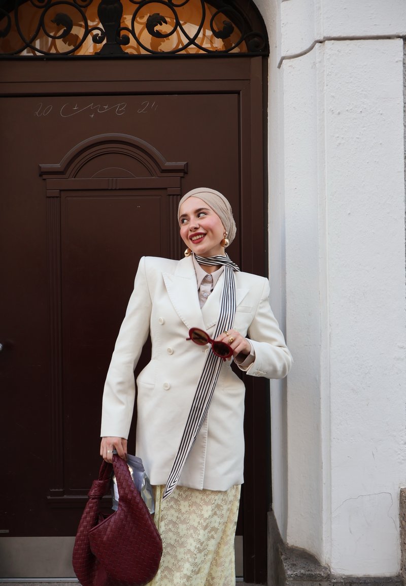 Blazer blanc à double boutonnage, écharpe rayée, hijab beige. Tenant des lunettes de soleil rouges et un sac tissé bordeaux. Jupe en dentelle visible sous le blazer.