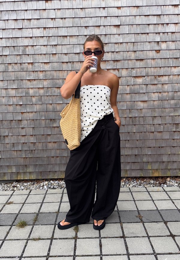 Top bianco senza spalline con pois neri, pantaloni neri a gamba larga, borsa a tracolla intrecciata color beige, infradito neri e occhiali da sole. Tiene una tazza di caffè.