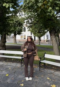 Brązowa skórzana kurtka, dopasowany top i rozkloszowane spodnie w tym samym kolorze, białe trampki, trzyma filiżankę kawy, otoczona drzewami i ławkami.