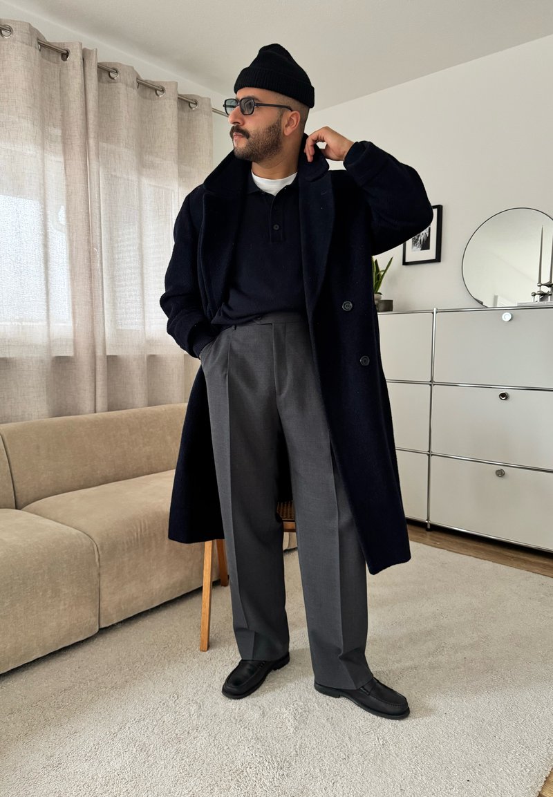 Man in black beanie, sunglasses, long black coat, navy shirt, gray trousers, and black shoes standing in modern living room.