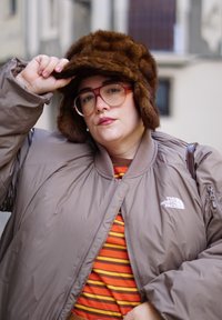 Άτομο με μεγάλα γυαλιά, καφέ γούνινο καπέλο, καφέ μπουφάν με πούπουλα και πορτοκαλί ριγέ πουκάμισο, που προσαρμόζει το καπέλο του σε εξωτερικό χώρο.