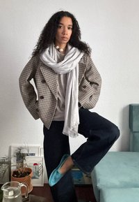 Rutete dobbeltradet blazer med store knapper, grå skjerf, beige topp, vide mørke jeans og turkise tøfler. Krøllete hår og nøytral bakgrunn.