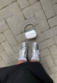 Sølv sneakers med strukturerte paneler og hvite snørebånd er parret med en liten metallisk veske. Bakgrunnen har grå steinfliser.