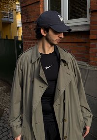 Overdimensjonert olivengrønn trenchcoat med store knapper, brukt over en tettsittende svart Nike-skjorte. En marineblå caps med LA-logo.