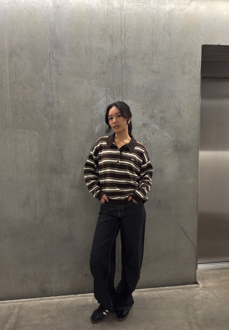 Polo a maniche lunghe a righe marroni e crema con colletto, abbinato a jeans a gamba larga scuri e sneakers nere, in piedi contro un muro grigio.