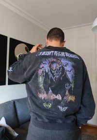 Inimene, kes kannab musta dressipluusi värvilise kolju graafika ja tekstiga seljal, seisab elutoas halli diivani lähedal.