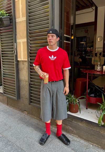Camisa roja de manga corta con acentos blancos y logo, pantalones cortos grises con pliegues, calcetines rojos y zuecos negros; sosteniendo un vaso de bebida fría.