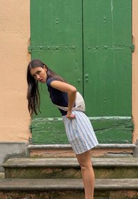 Modelo vestida con una blusa ajustada de color azul oscuro y una falda de rayas verticales en azul claro. Lleva una riñonera beige y está de pie en unos escalones de piedra cerca de puertas verdes.