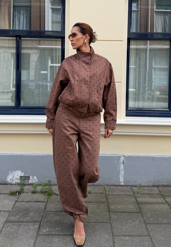 Femme en veste et pantalon marron à pois, chaussures beiges, lunettes de soleil et cheveux en chignon, debout sur un trottoir de ville devant des fenêtres.