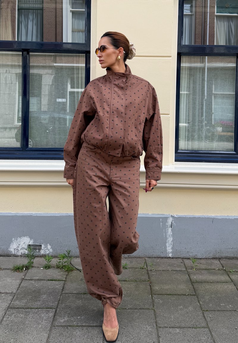 Woman in brown polka dot jacket and pants, beige shoes, sunglasses, and hair in a bun standing on a city sidewalk in front of windows.