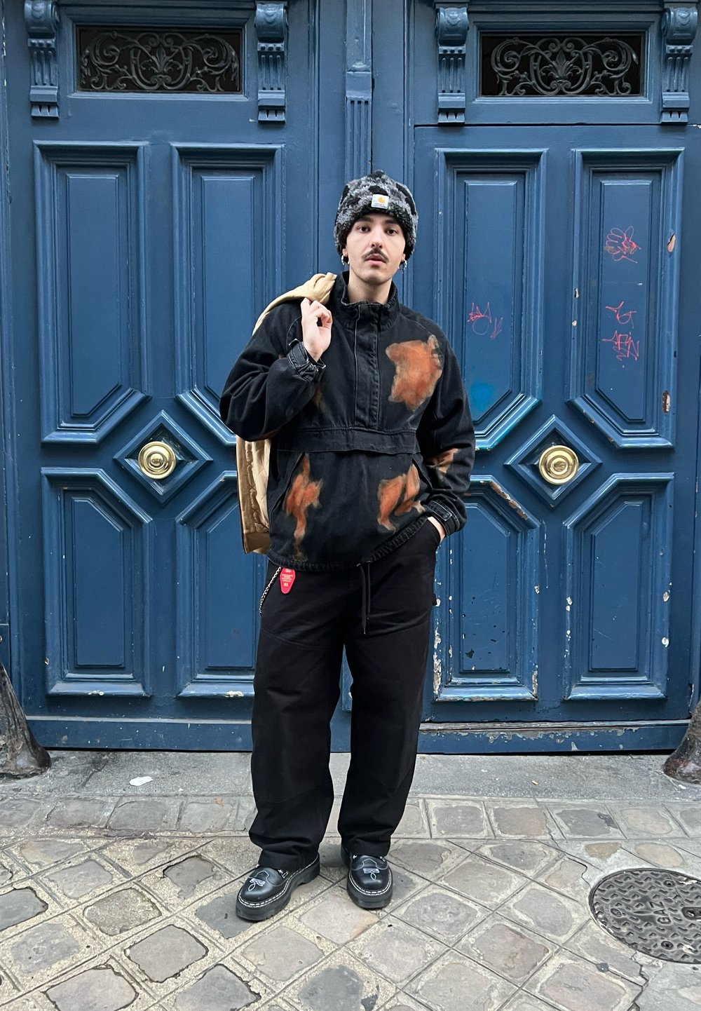 Black cropped jacket with orange stains, black trousers, and black shoes. Beige backpack slung over shoulder. Background features blue doors.