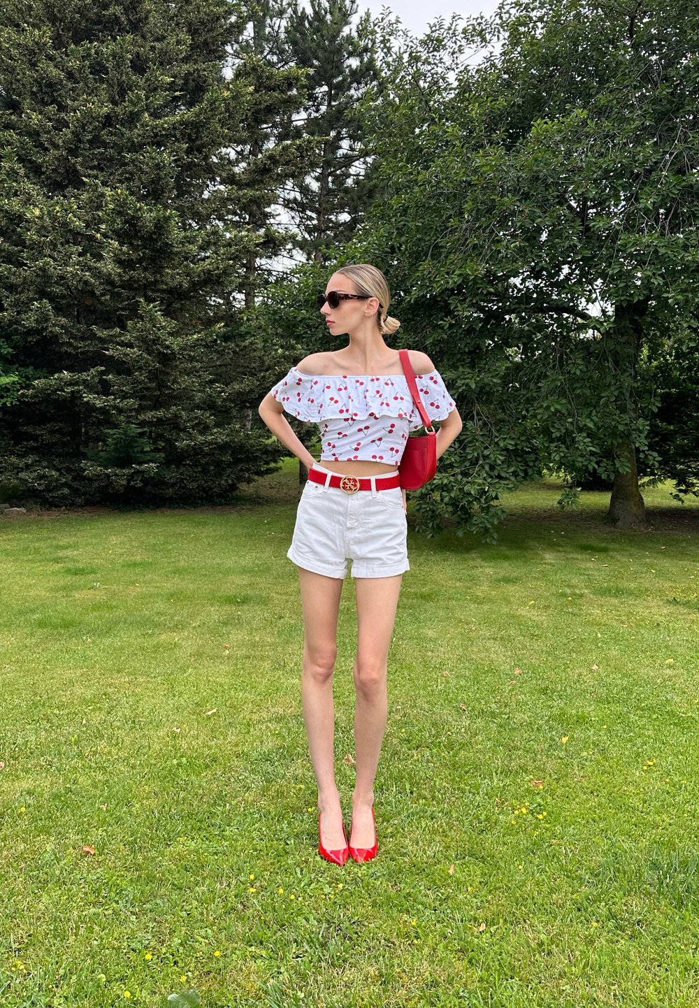 Top bianco scollato con stampa di ciliegie rosse, abbinato a pantaloni corti bianchi, cintura rossa e tacchi alti rossi; porta una borsa a tracolla rossa.