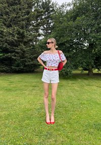 Top bianco scollato con stampa di ciliegie rosse, abbinato a pantaloni corti bianchi, cintura rossa e tacchi alti rossi; porta una borsa a tracolla rossa.