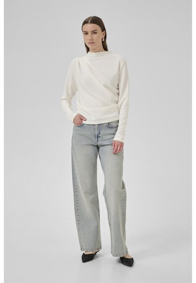 Top de manga larga crema con diseño drapeado, combinado con jeans de pierna ancha en azul claro y zapatos planos negros de punta. Textura de tela simple y suave.